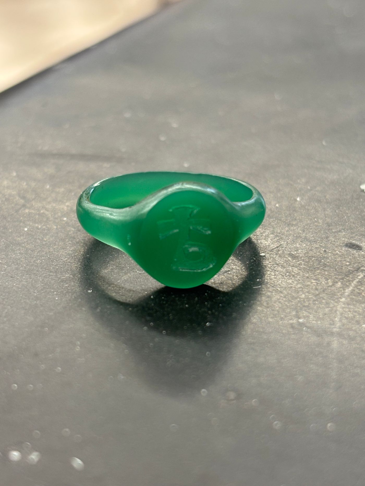 Modèle en cire verte d'une chevalière, plateau ovale gravé d'un caractère, posé sur une surface noire.
