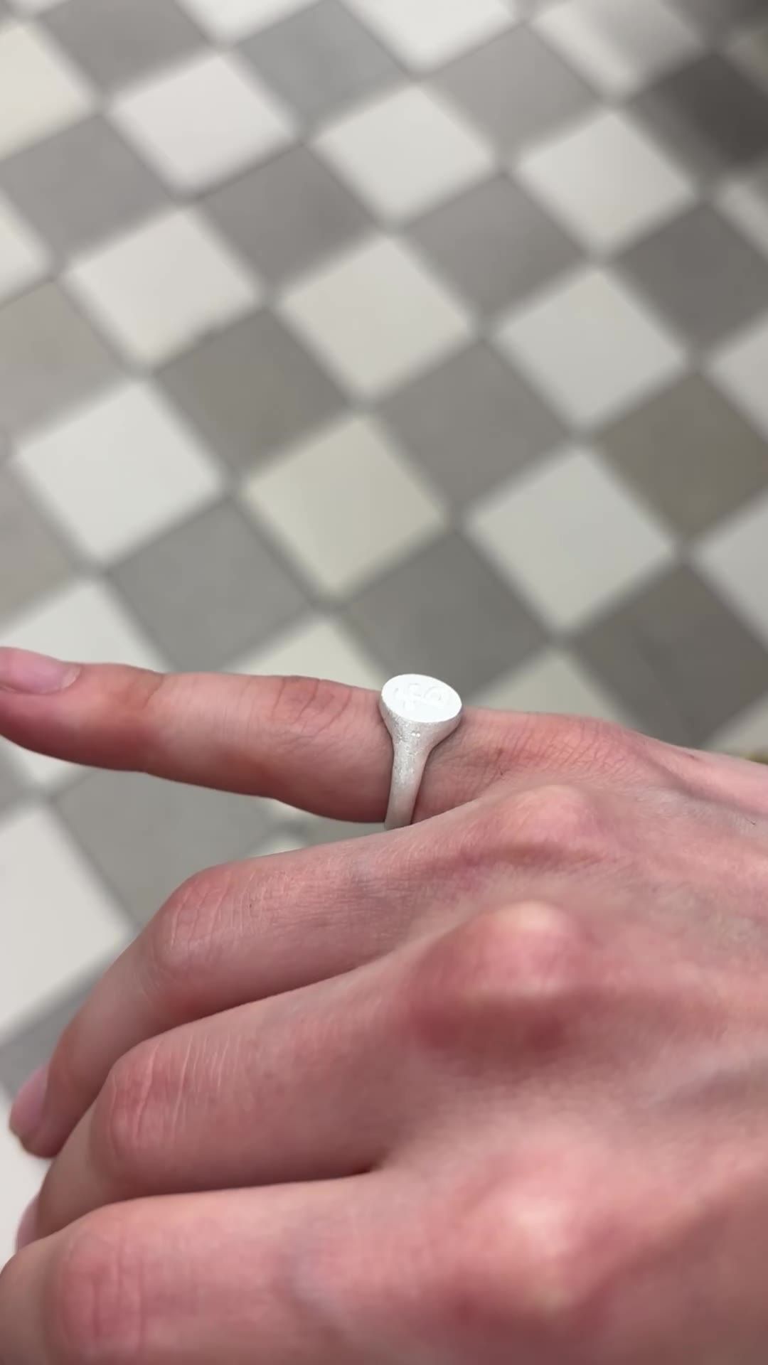 Chevalière en argent portée à l'index, plateau ovale gravé, au-dessus d'un sol en damier (image tirée du clip).