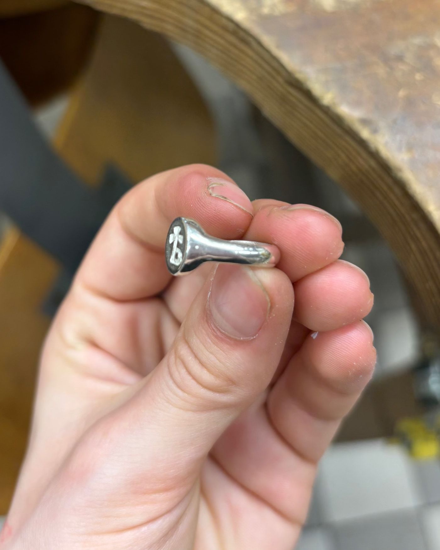 Chevalière en argent tenue entre les doigts, de trois quarts, épaulement poli qui s'évase vers le plateau ovale gravé, cheville d'établi en bois derrière.