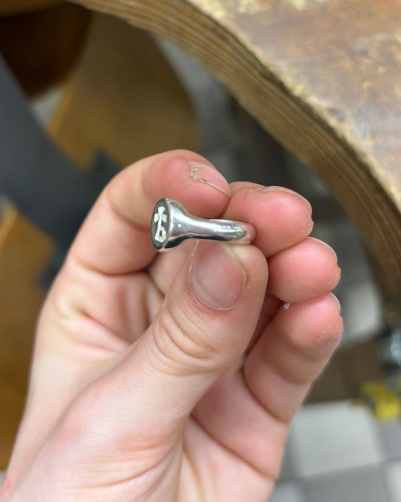 Chevalière en argent tenue entre les doigts, de trois quarts, épaulement poli qui s'évase vers le plateau ovale gravé, cheville d'établi en bois derrière.