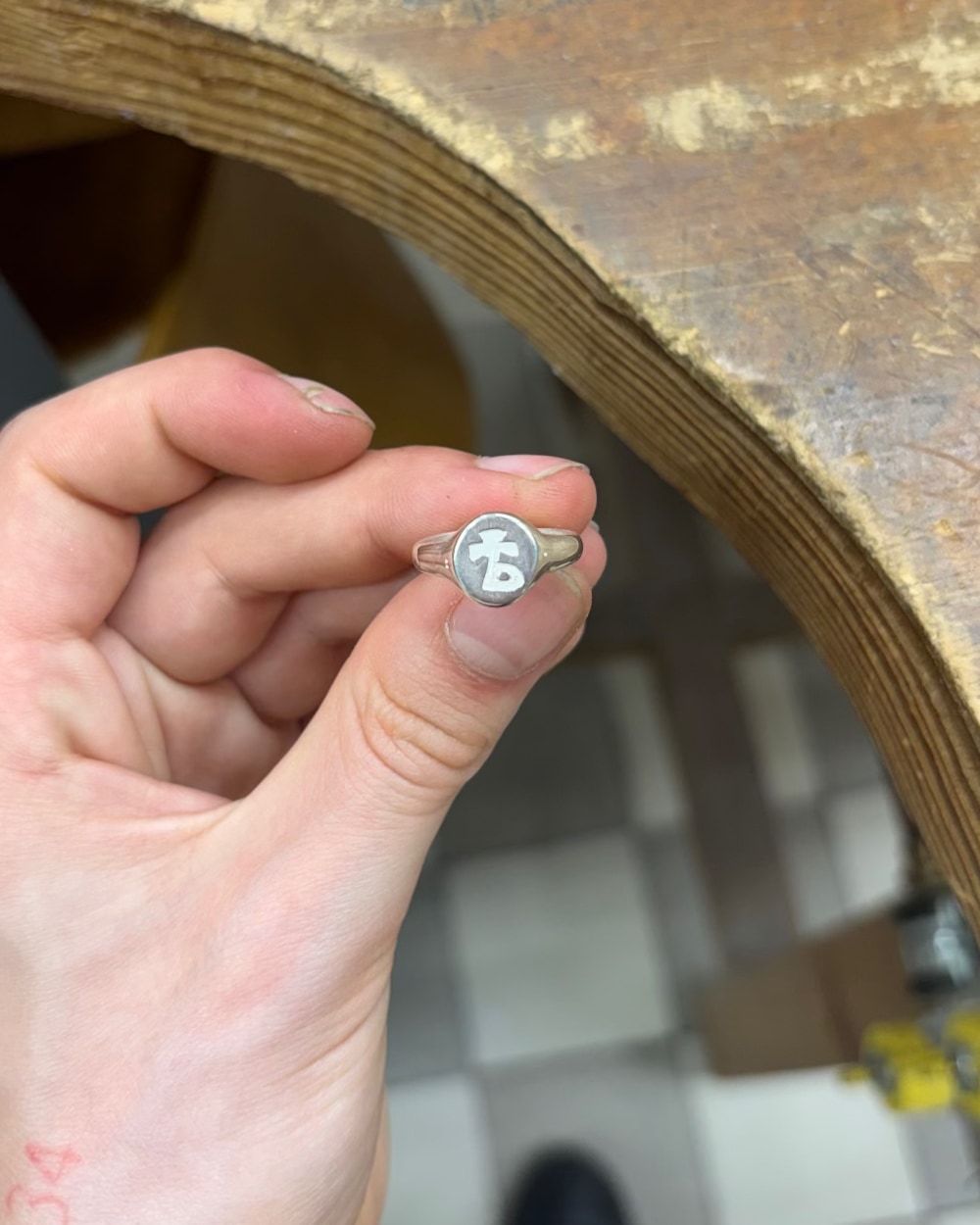 Chevalière en argent vue de face, caractère gravé en réserve sur le plateau ovale mat, la bague pincée entre le pouce et l'index au-dessus de l'établi.