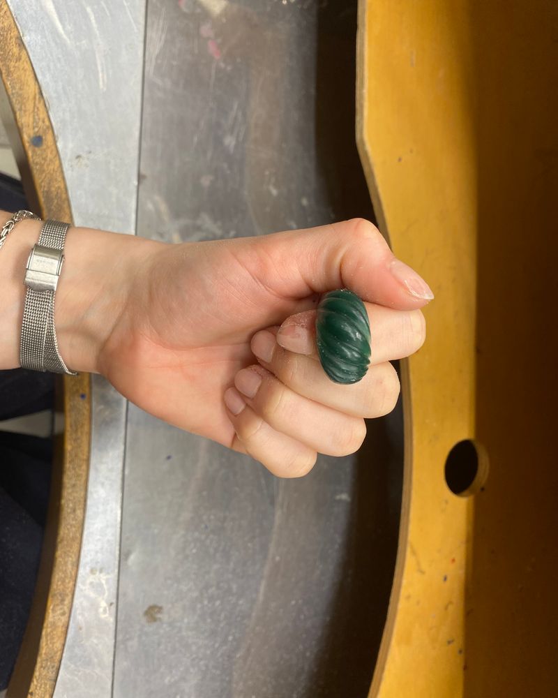 Modèle en cire verte d'une bague bombée à cannelures torsadées, tenu dans la main au-dessus de l'établi.