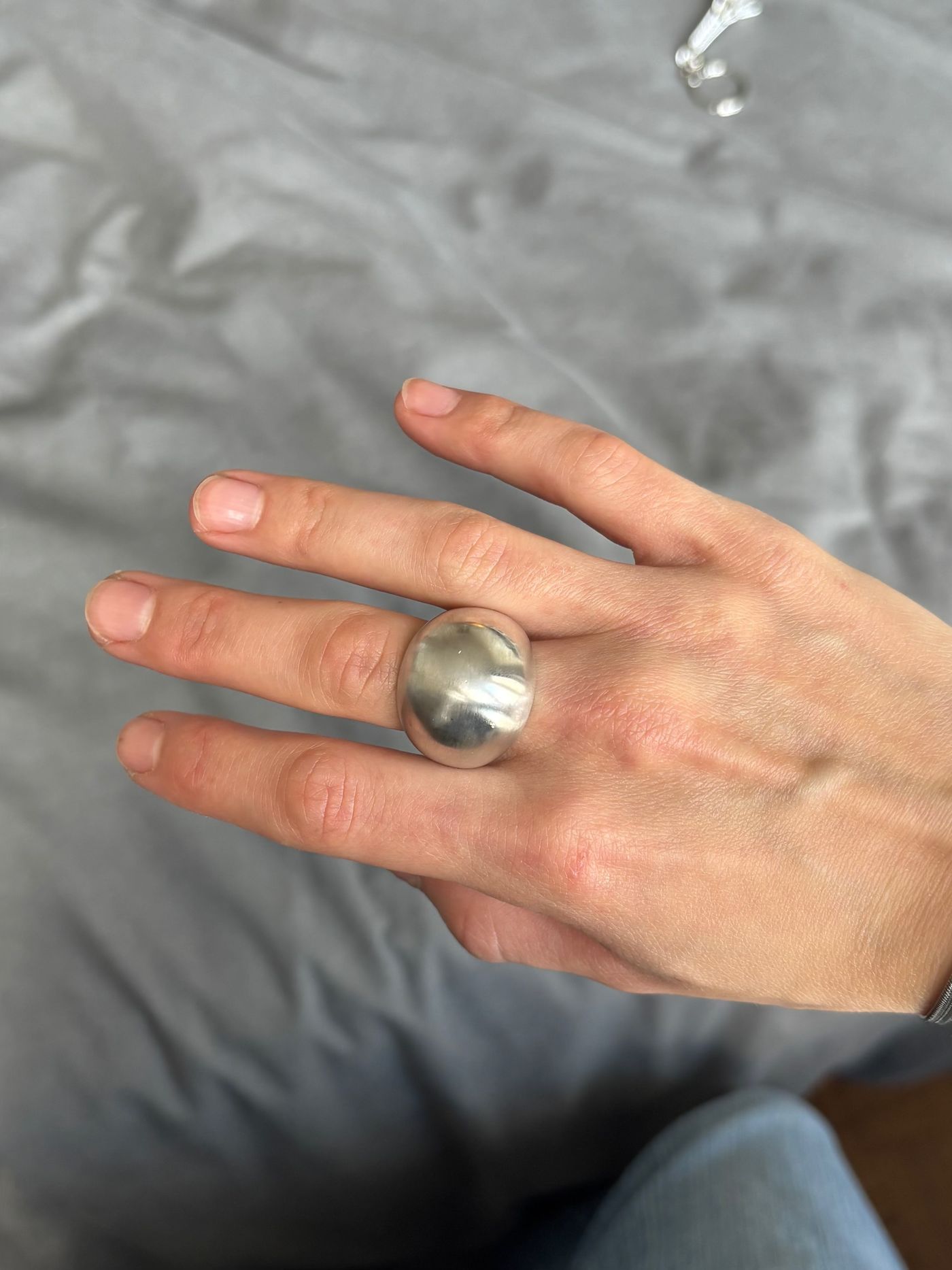 Bague en argent à large dôme lisse, portée au majeur, le poli miroir reflétant la main et le tissu gris derrière.