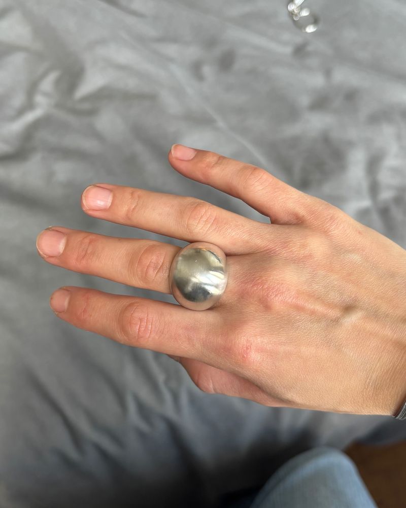 Bague en argent à large dôme lisse, portée au majeur, le poli miroir reflétant la main et le tissu gris derrière.