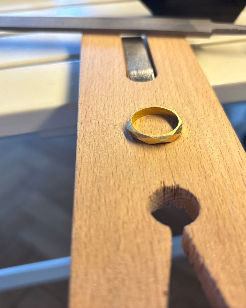 Anneau en laiton à facettes irrégulières, posé sur une cheville d'établi en bois, lime en arrière-plan.