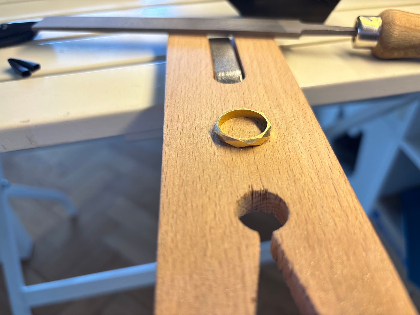 Anneau en laiton à facettes irrégulières, posé sur une cheville d'établi en bois, lime en arrière-plan.