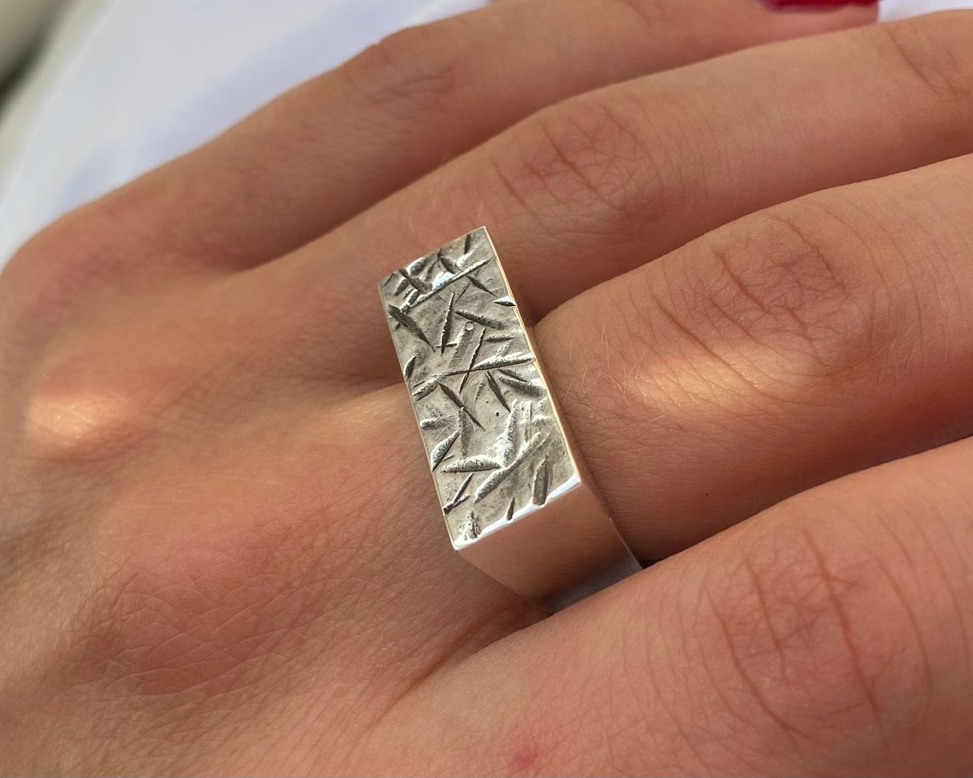 Bague portée, vue de côté, plateau rectangulaire texturé et anneau lisse, les deux mains posées sur tissu blanc.