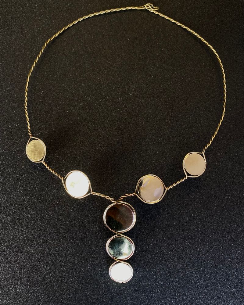 Collier en fil de laiton torsadé portant sept disques, quatre sur le tour de cou et trois en pendant vertical, sur fond noir.