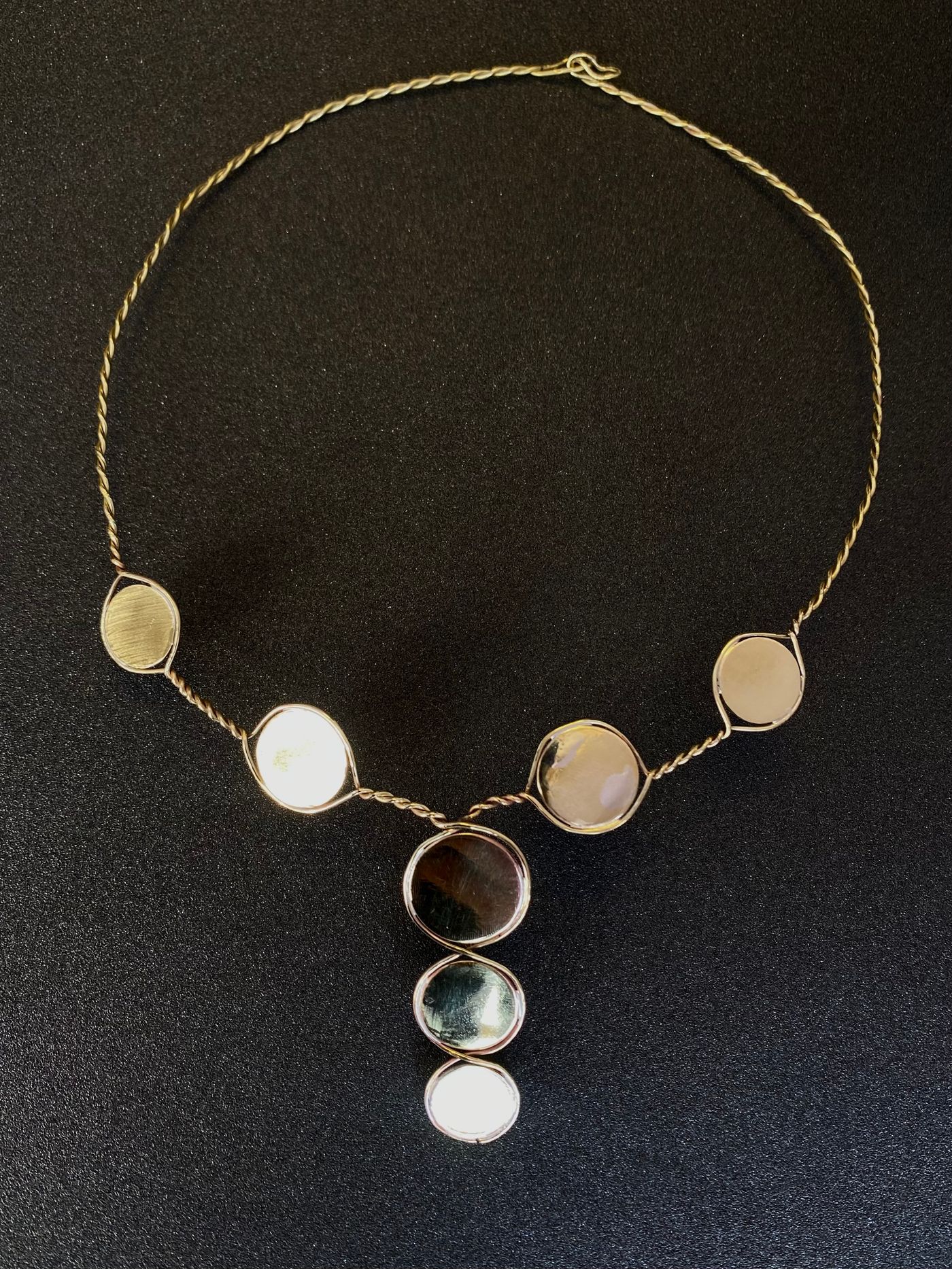 Collier en fil de laiton torsadé portant sept disques, quatre sur le tour de cou et trois en pendant vertical, sur fond noir.
