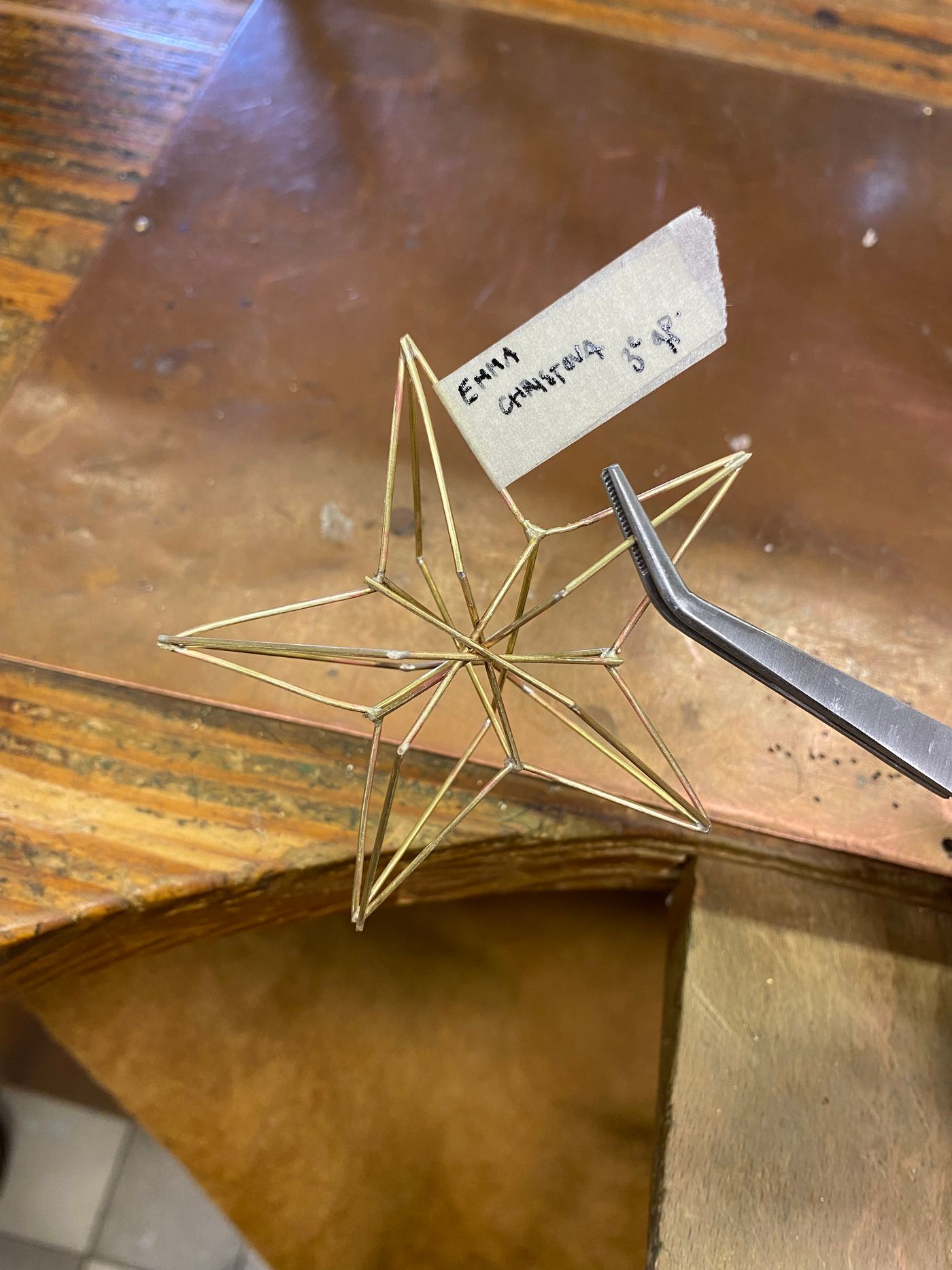 Étoile à cinq branches en fil de laiton, structure en trois dimensions, tenue à la pince sur l'établi, étiquette de ruban au nom d'Emma Christova.