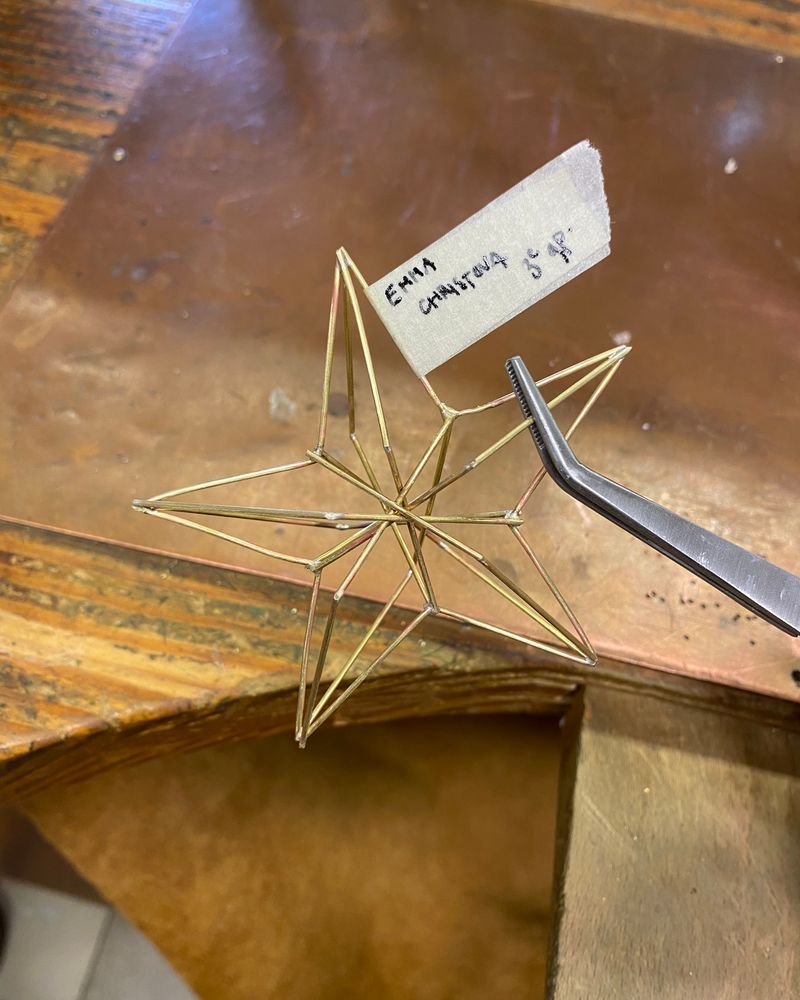Étoile à cinq branches en fil de laiton, structure en trois dimensions, tenue à la pince sur l'établi, étiquette de ruban au nom d'Emma Christova.